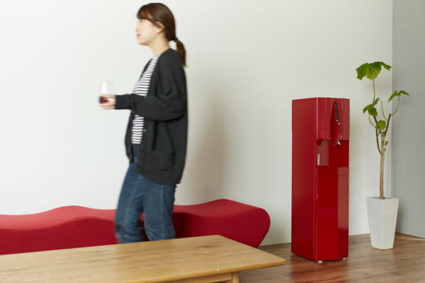 テーブルとソファーのある部屋に設置されている赤いウォーターサーバーとグラスを持って歩く女性
